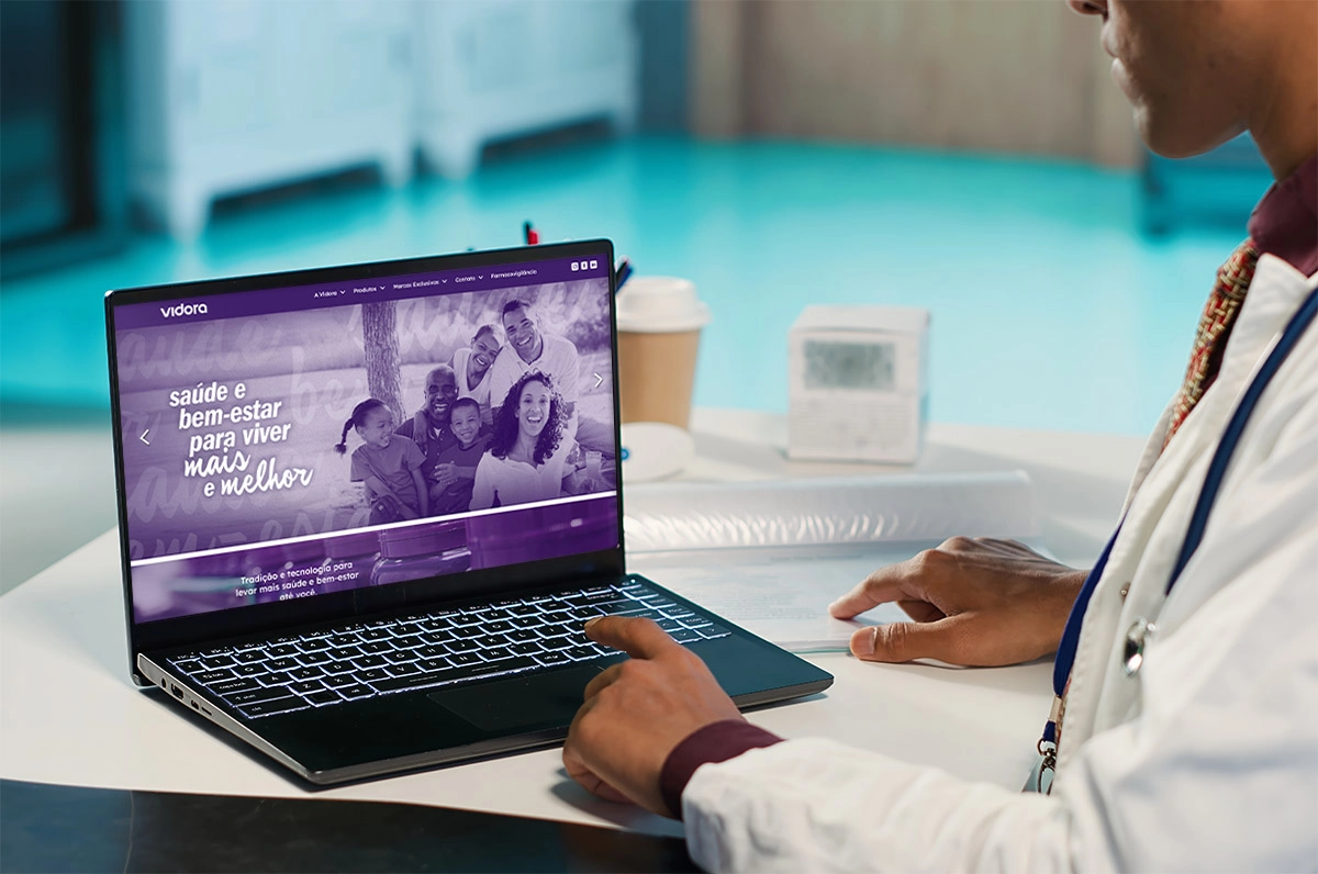 A doctor interacting with a notebook screen displaying the homepage of the Vidora website, which uses a dominant purple color scheme. The main banner features a photograph of a smiling multi-generational family in a park setting overlaid with the text 'Health and well-being to live longer and better'.