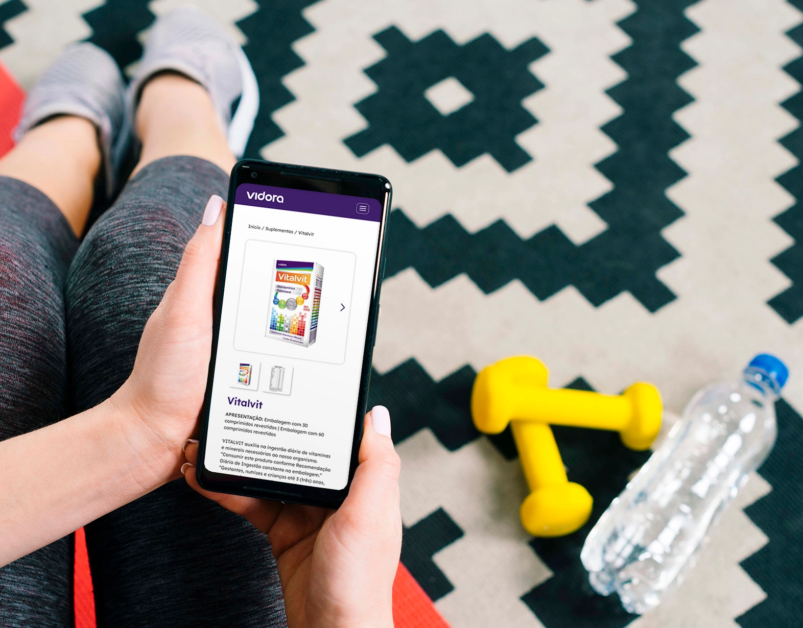 A person holding a smartphone displaying the product page for Vitalvit on the Vidora website. The page shows an image of the Vitalvit box, navigation breadcrumbs, and a detailed text description of the product. Next to the person's feet are a clear bottle of water and two bright
          yellow dumbbells.
